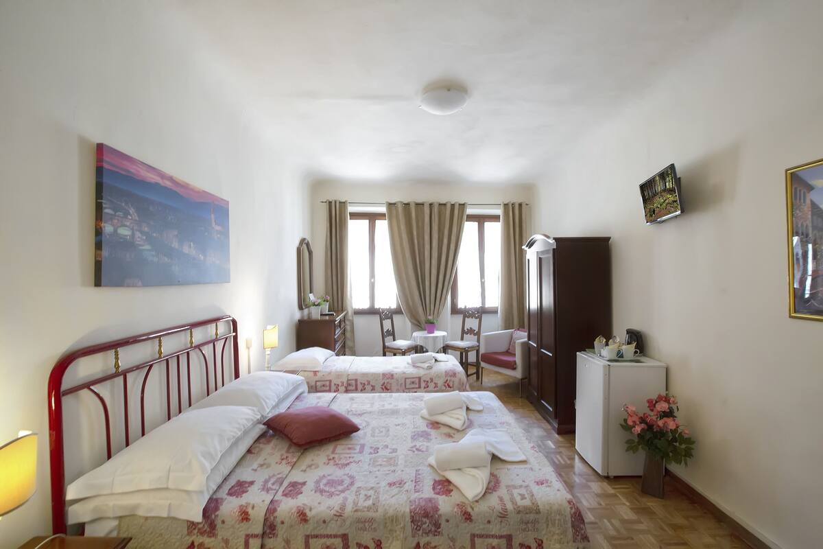A hotel room with two beds, a desk, a chair, a small table, a refrigerator, and a window with curtains.