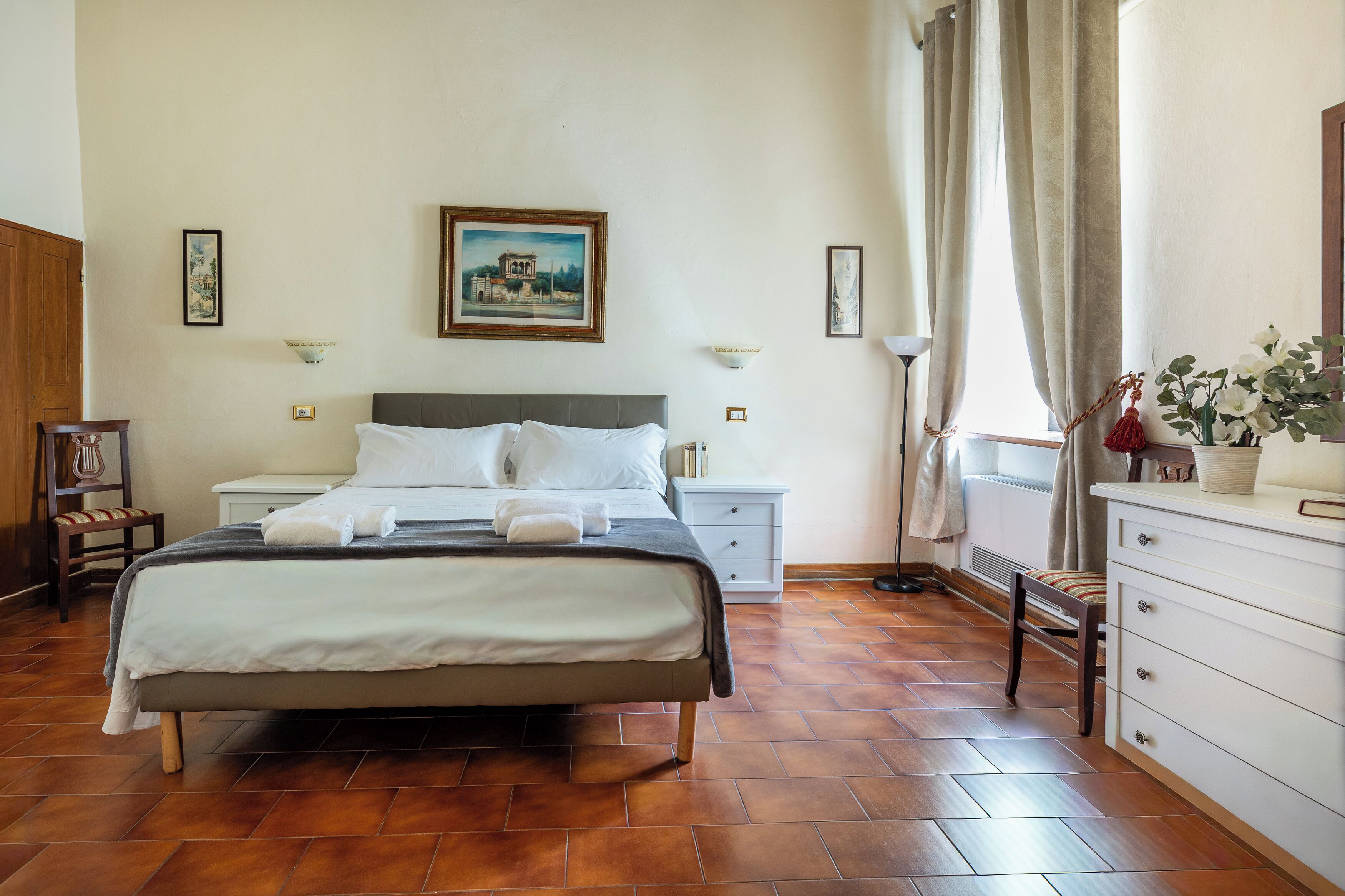 Une chambre d’hôtel bien rangée, avec un lit, des tables de chevet, une chaise, une commode et une fenêtre avec des rideaux.
