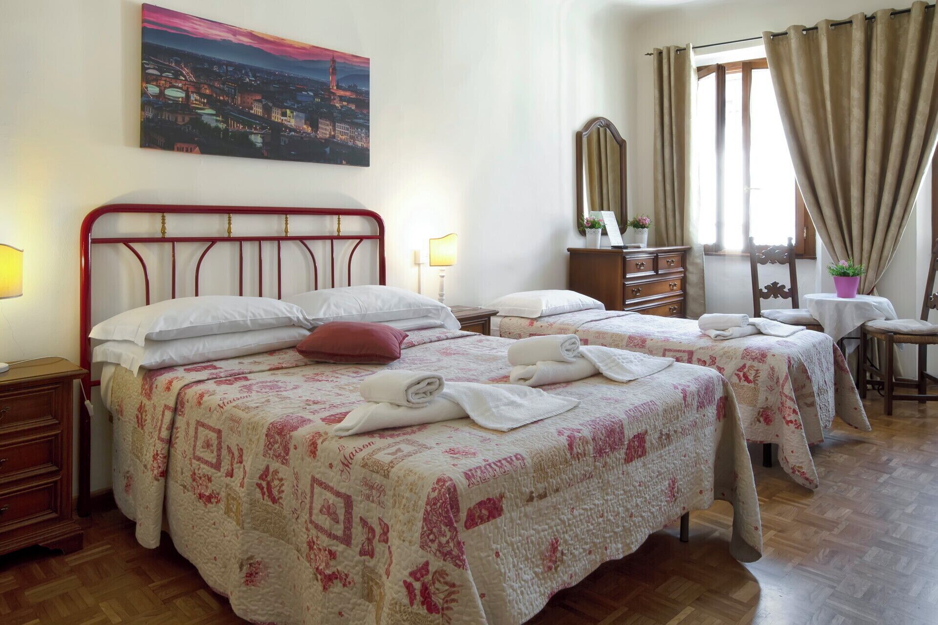 Une chambre d’hôtel avec deux lits, une commode en bois, un bureau avec une chaise et un tableau représentant un paysage urbain accroché au mur.