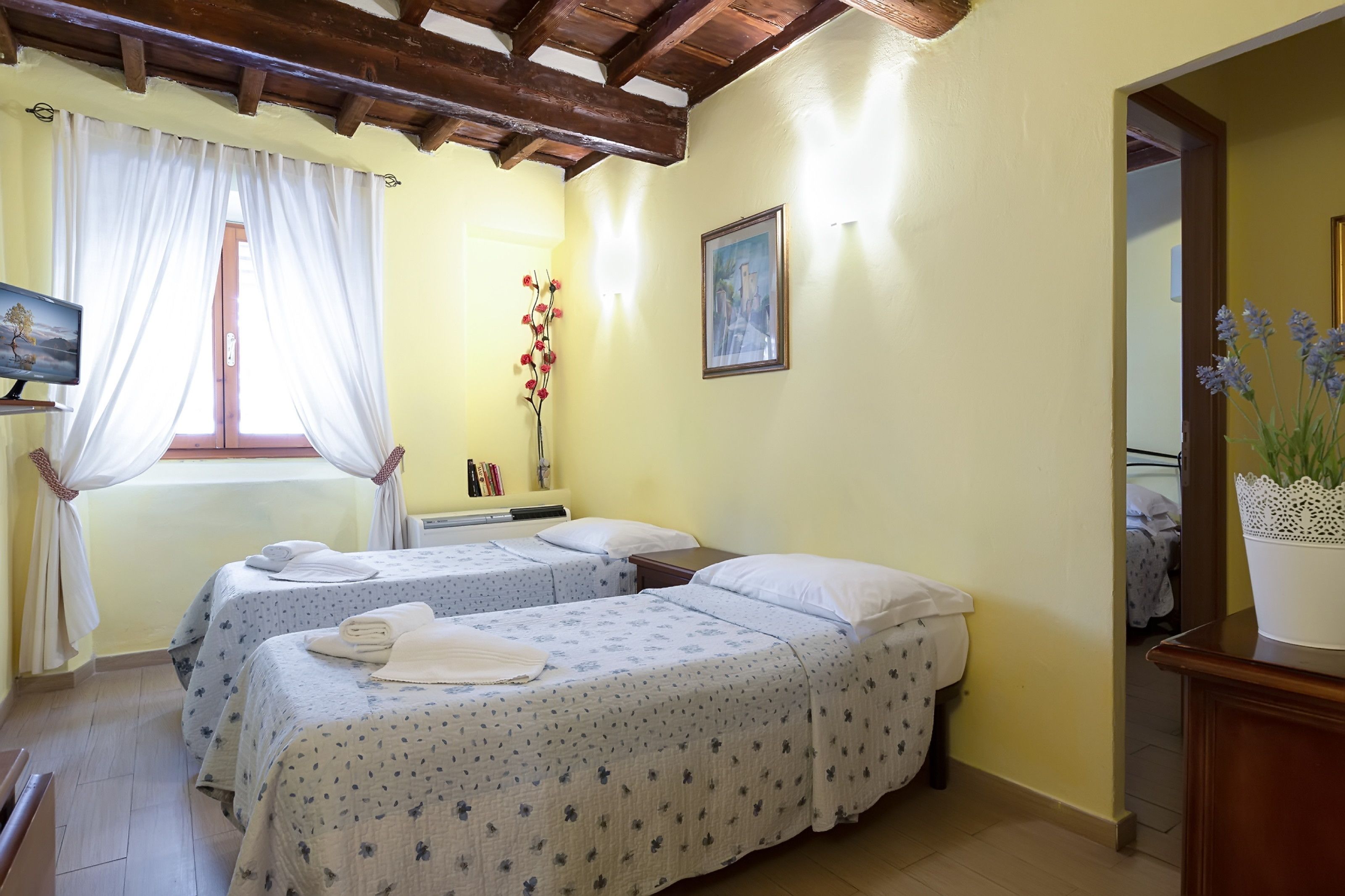 A room with two beds, a wooden ceiling, a window with white curtains, a painting on the wall, and a vase with flowers.