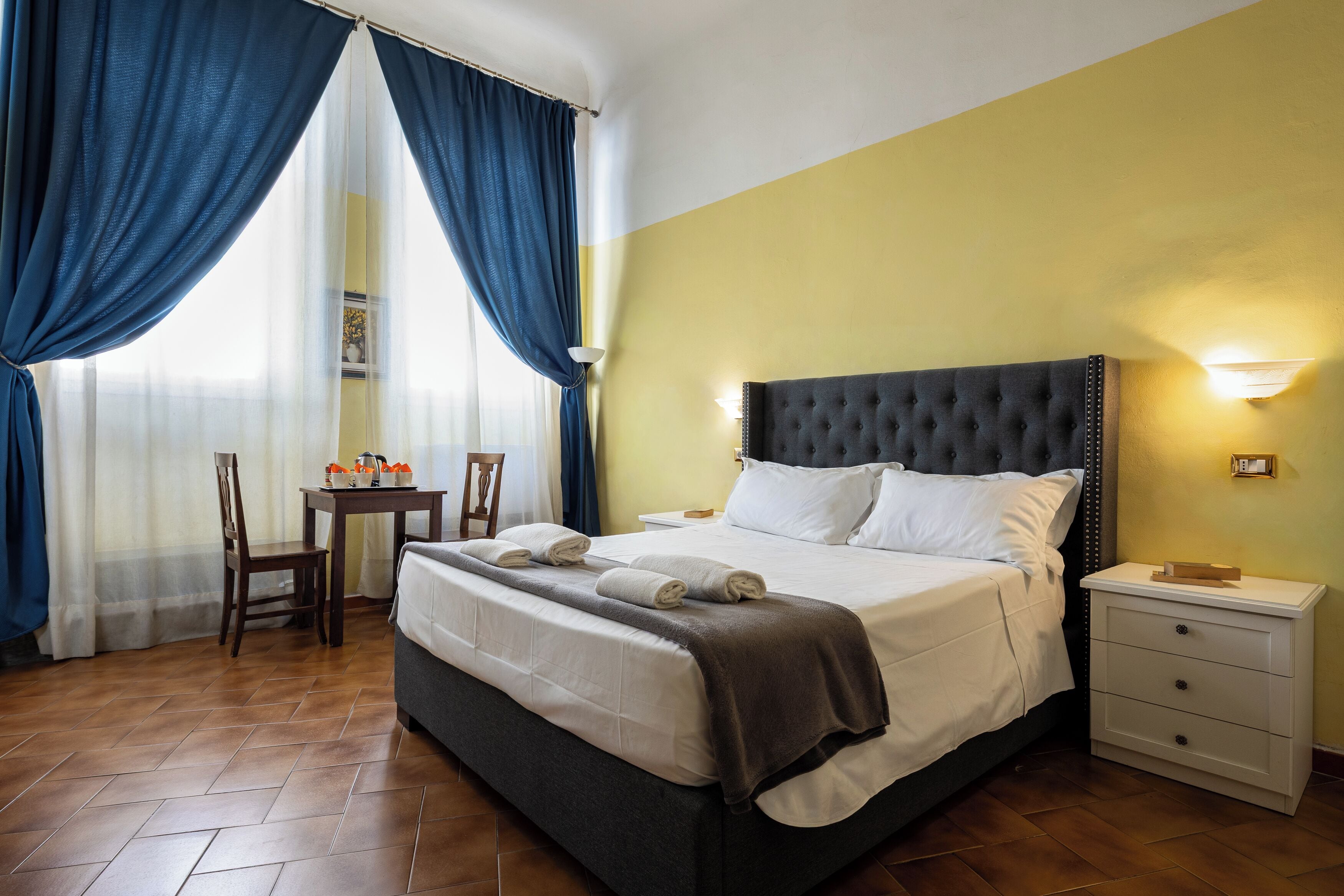 A hotel room with a bed, a nightstand, a chair, a table, and curtains.