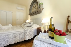 Quarto de hotel com duas camas, uma mesinha de cabeceira, uma mesa com uma bandeja de champanhe e morangos, e um objeto decorativo na parede acima da cama.