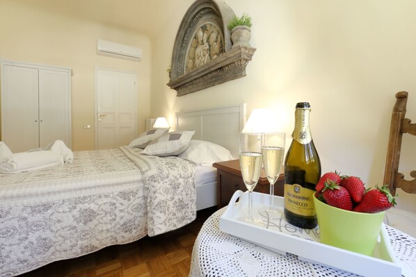A hotel room with two beds, a nightstand, a table with a tray of champagne and strawberries, and a decorative wall piece above the bed.