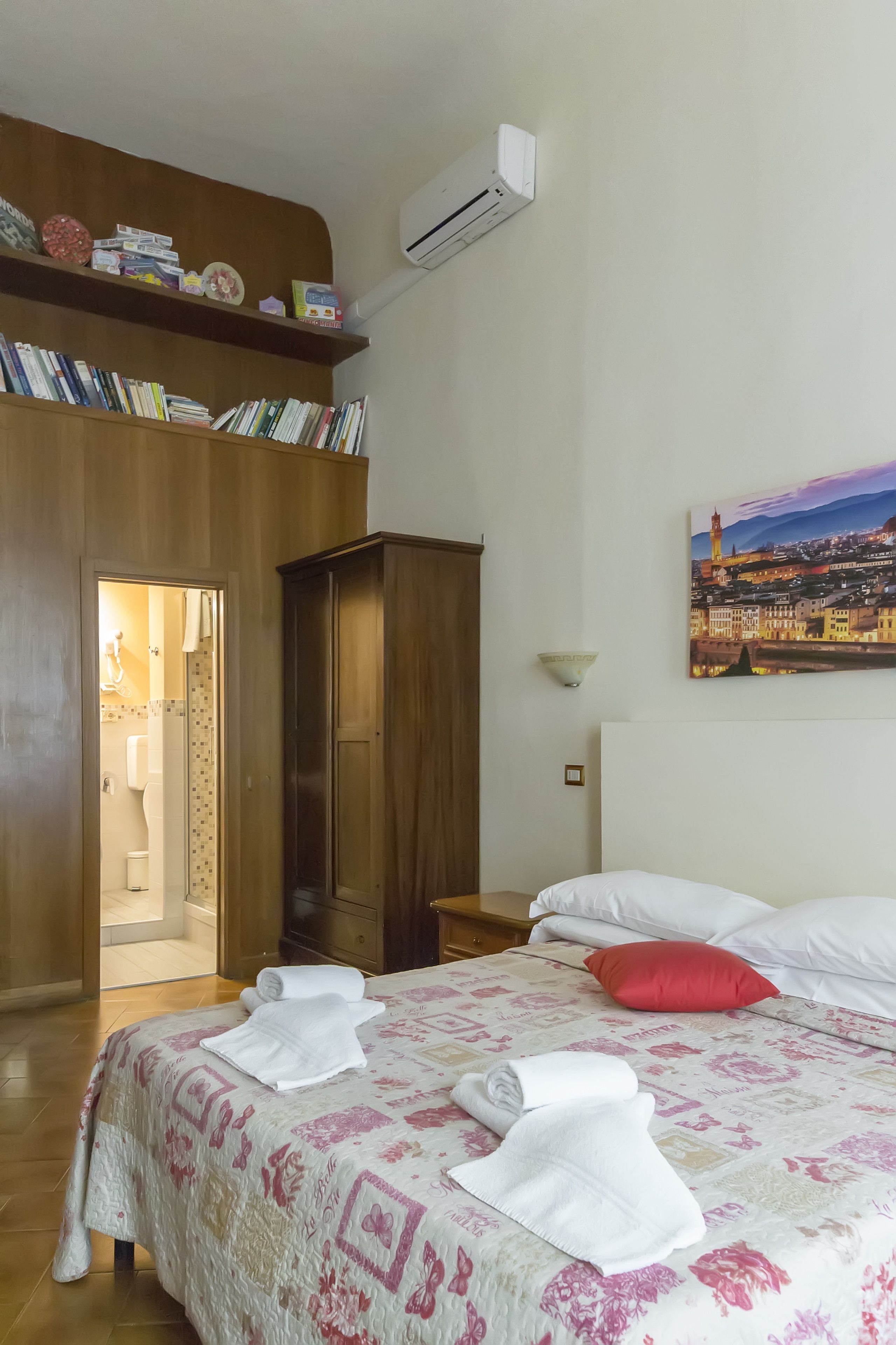 A hotel room with a bed, a wooden wardrobe, a bookshelf, a painting, and an air conditioning unit.
