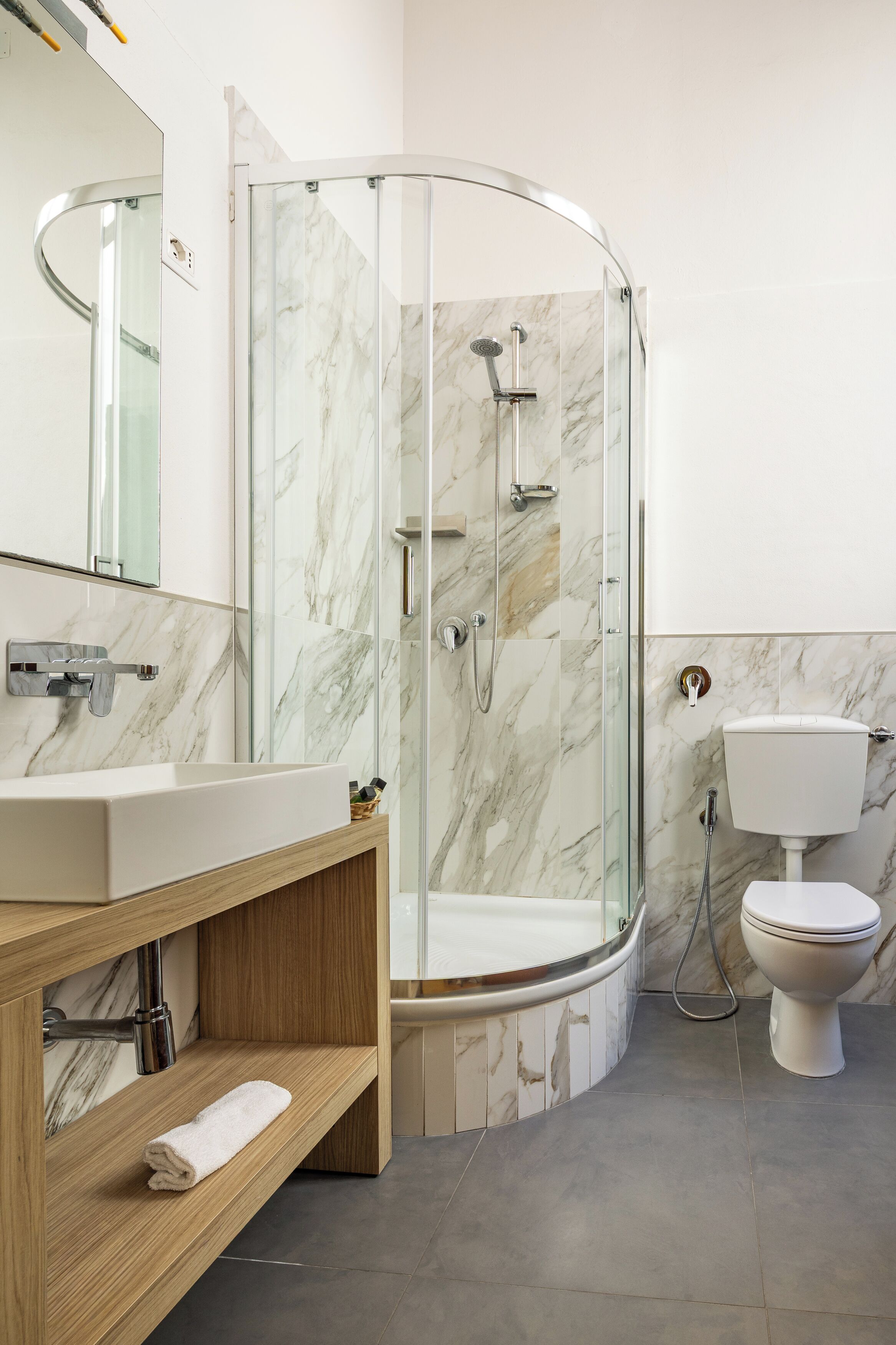 Une salle de bain moderne avec une douche à parois de verre, un mur recouvert de carreaux de marbre, des toilettes blanches et un meuble-lavabo en bois.