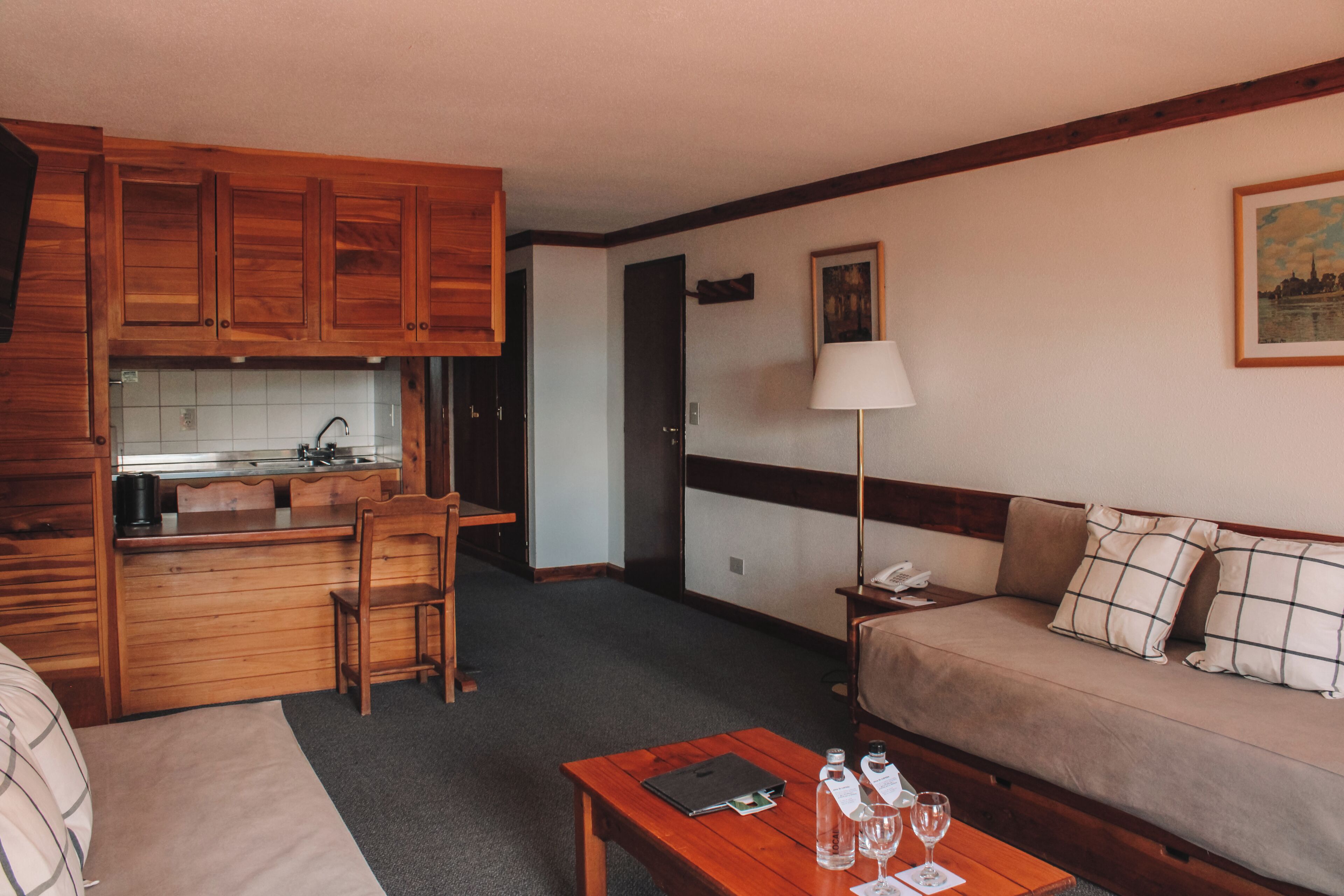 A compact hotel room with a kitchenette, a sofa, a coffee table, and a lamp.