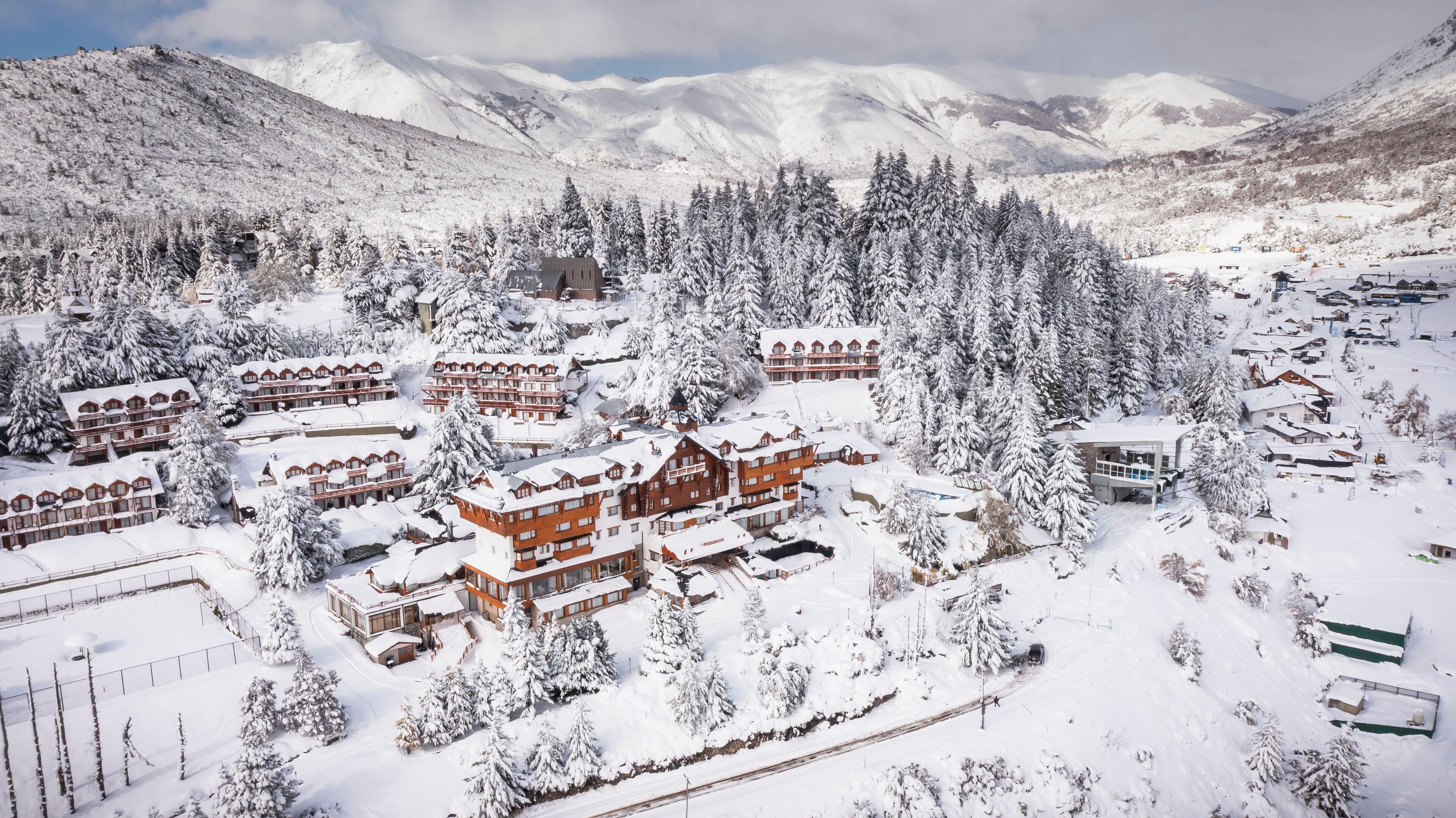 Un centro turístico de montaña cubierto de nieve, con edificios de estilo chalet, una pista de esquí y un fondo de bosque.