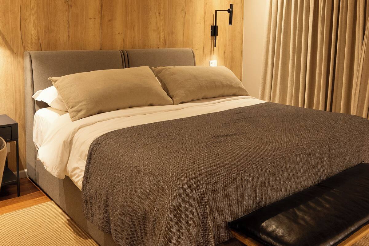 Ein modernes Schlafzimmer mit einem Holz-Kopfteil, einem Bett mit grau-weißer Bettwäsche, einer Bank am Fußende des Bettes und einem Deckenventilator.