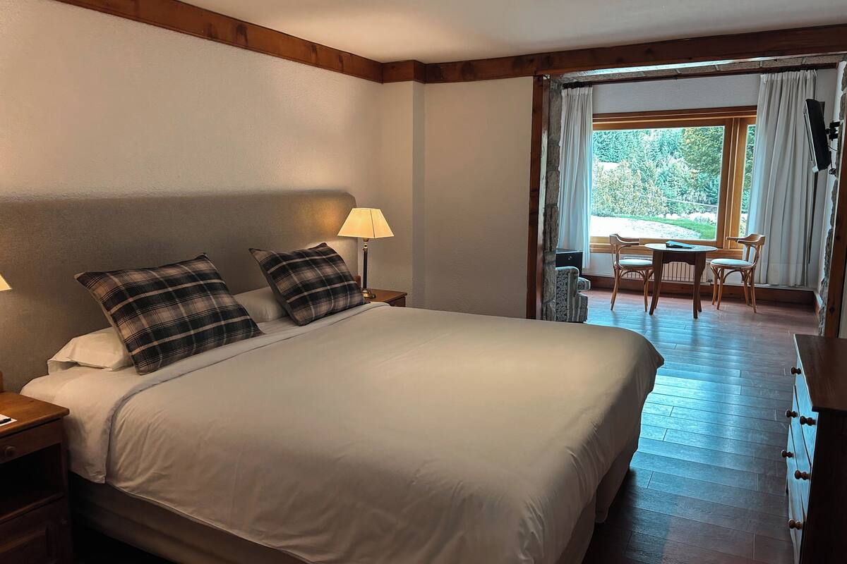 Ein Schlafzimmer mit einem großen Bett, zwei Nachttischlampen und Blick auf einen Außenbereich mit Essplatz durch ein Fenster.