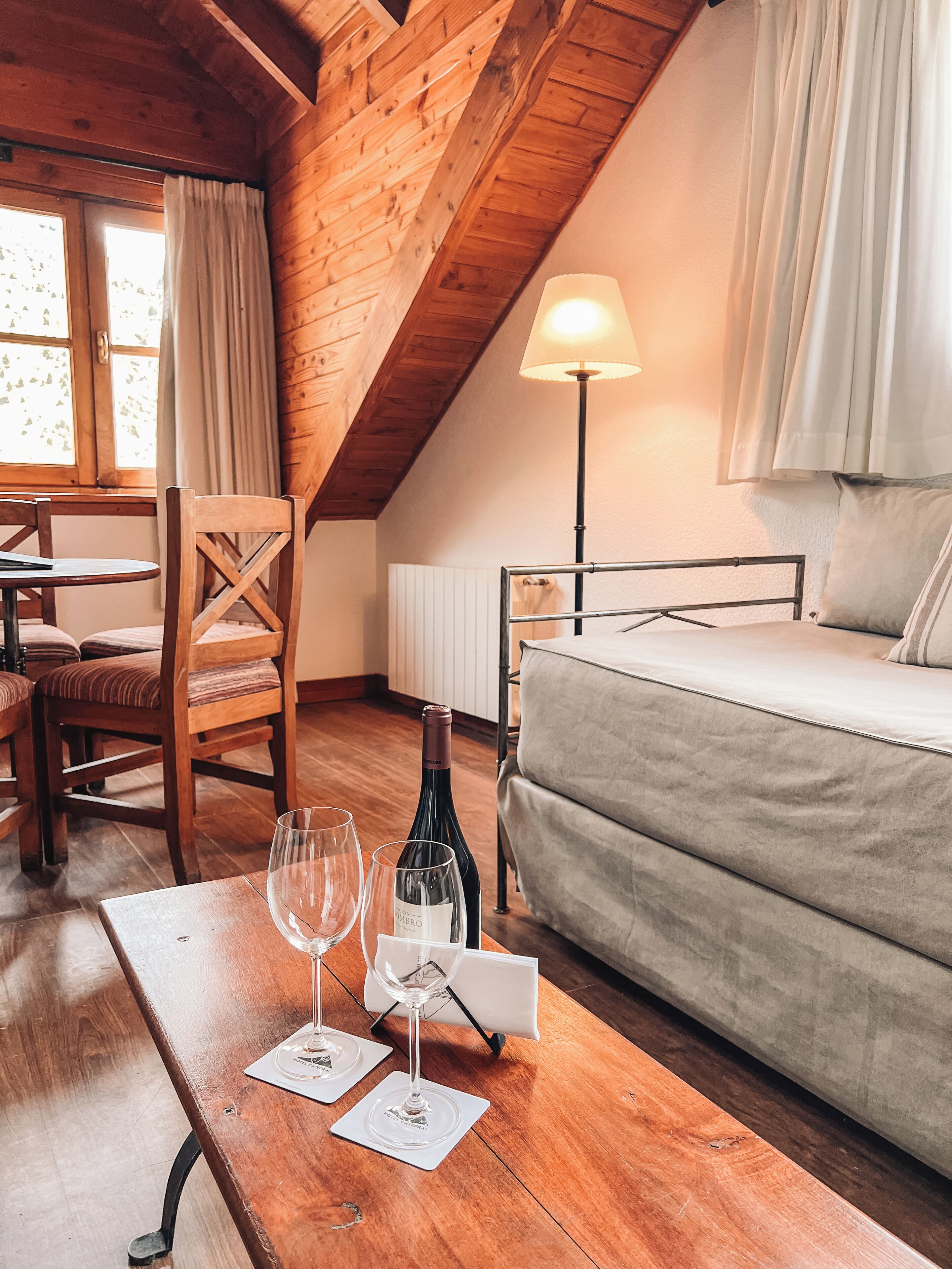A cozy room with wooden beams, a bed, a table with wine glasses, and a lamp.