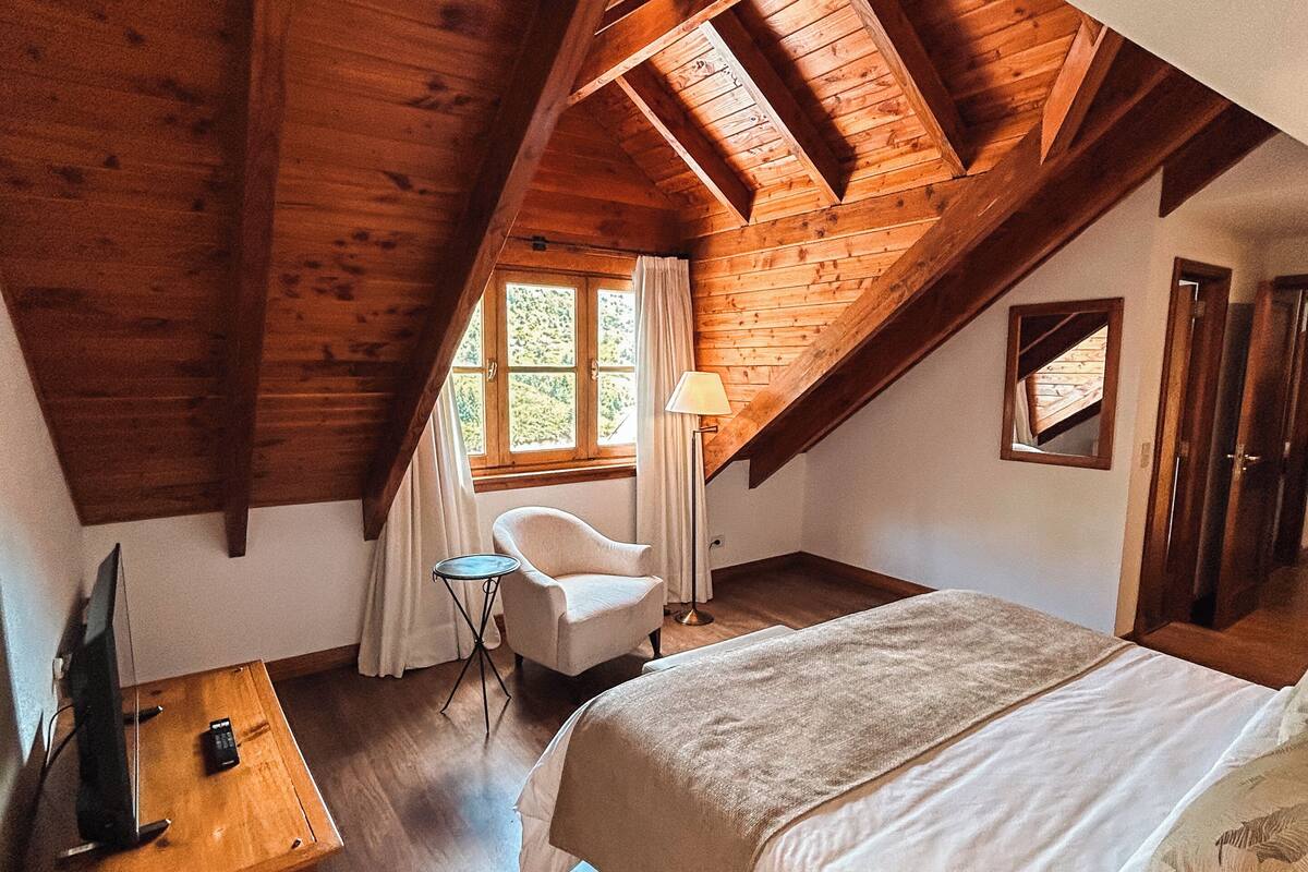 Ein Schlafzimmer mit schrägem Dach, einem Bett, einem Sessel, einem Schreibtisch mit Fernbedienung und einem Fenster mit Vorhängen.