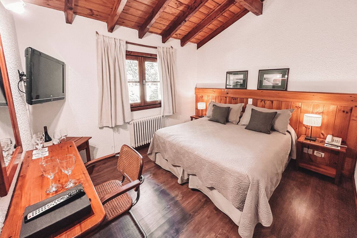 Ein Schlafzimmer mit Holzboden, einem Bett mit Kopfteil, einem an der Wand befestigten Fernseher, einem Fenster mit Vorhängen, einem Nachttisch mit Lampe und einem Sessel.