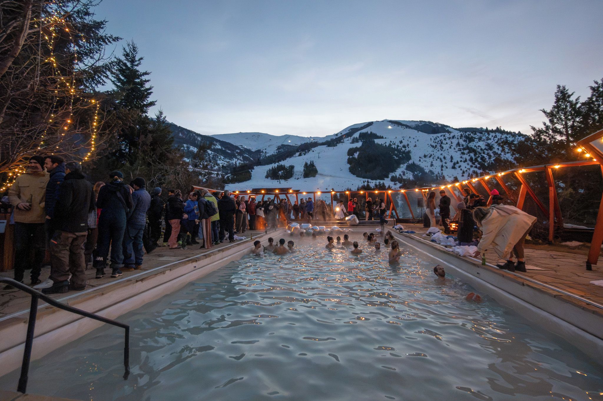 A swimming pool with people around it, surrounded by trees and mountains in the background.