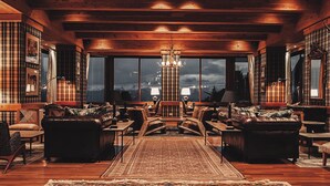 A cozy, log-furnished living room with a large window, a chandelier, and a central fireplace. - Hotel Catedral Patagonia Ski & Wellness (San Carlos de Bariloche)