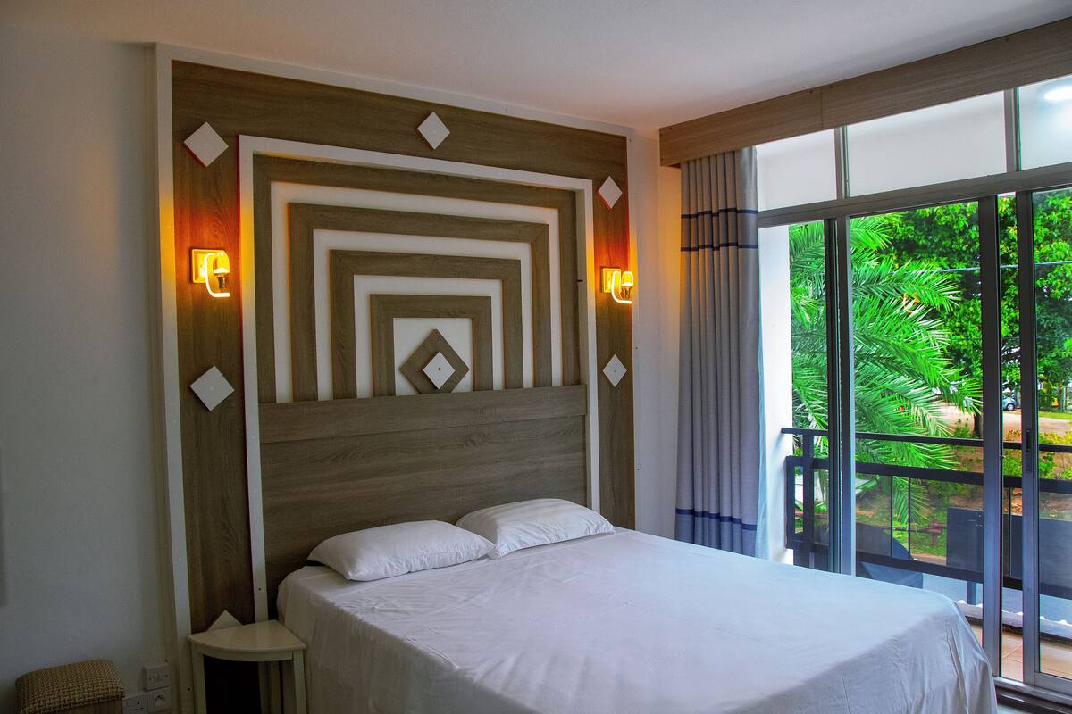 A hotel room with a large bed, two bedside lamps, and a view of greenery through the window.