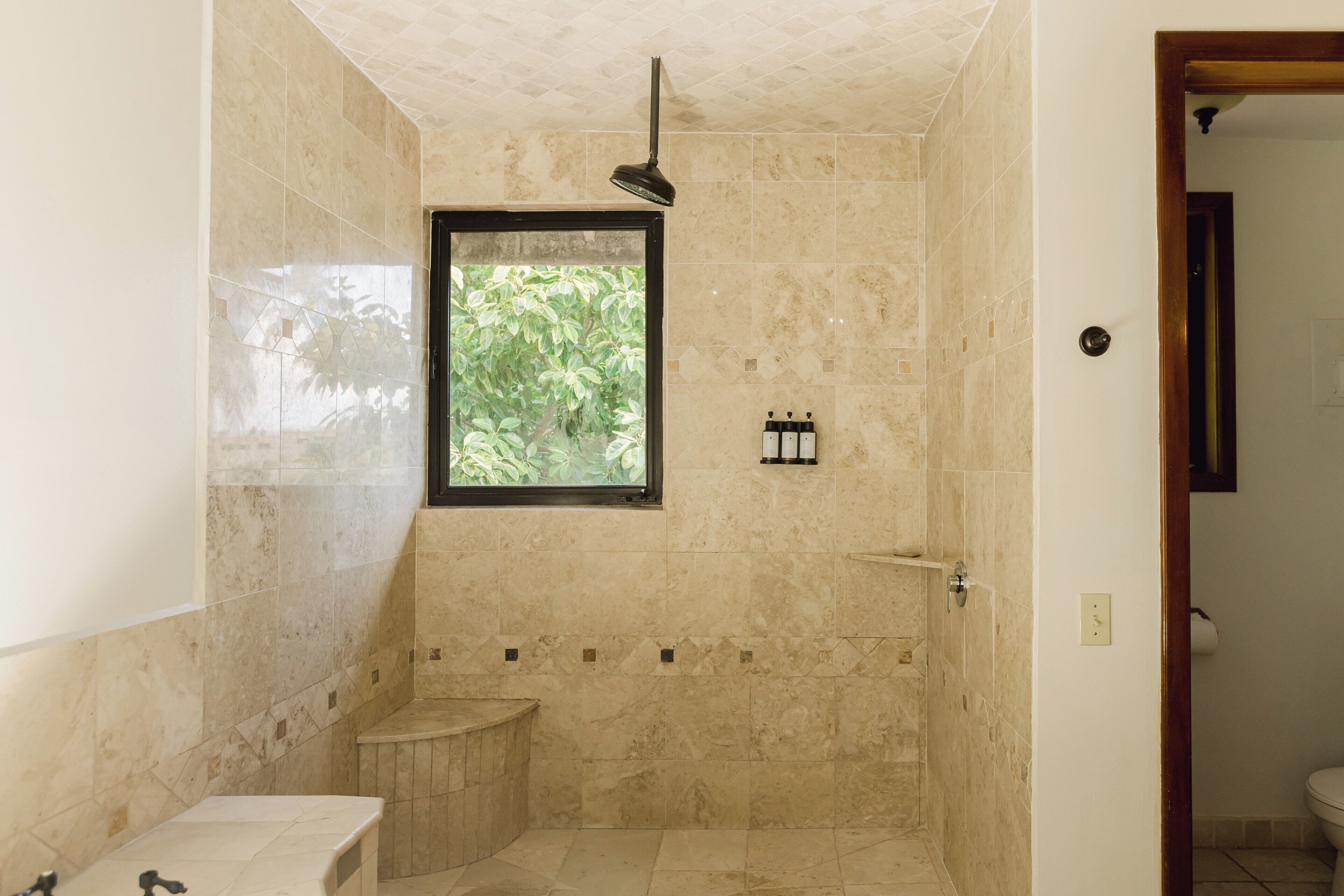 A bathroom with a shower, toilet, and sink, featuring beige tiles and a window with a view of greenery.