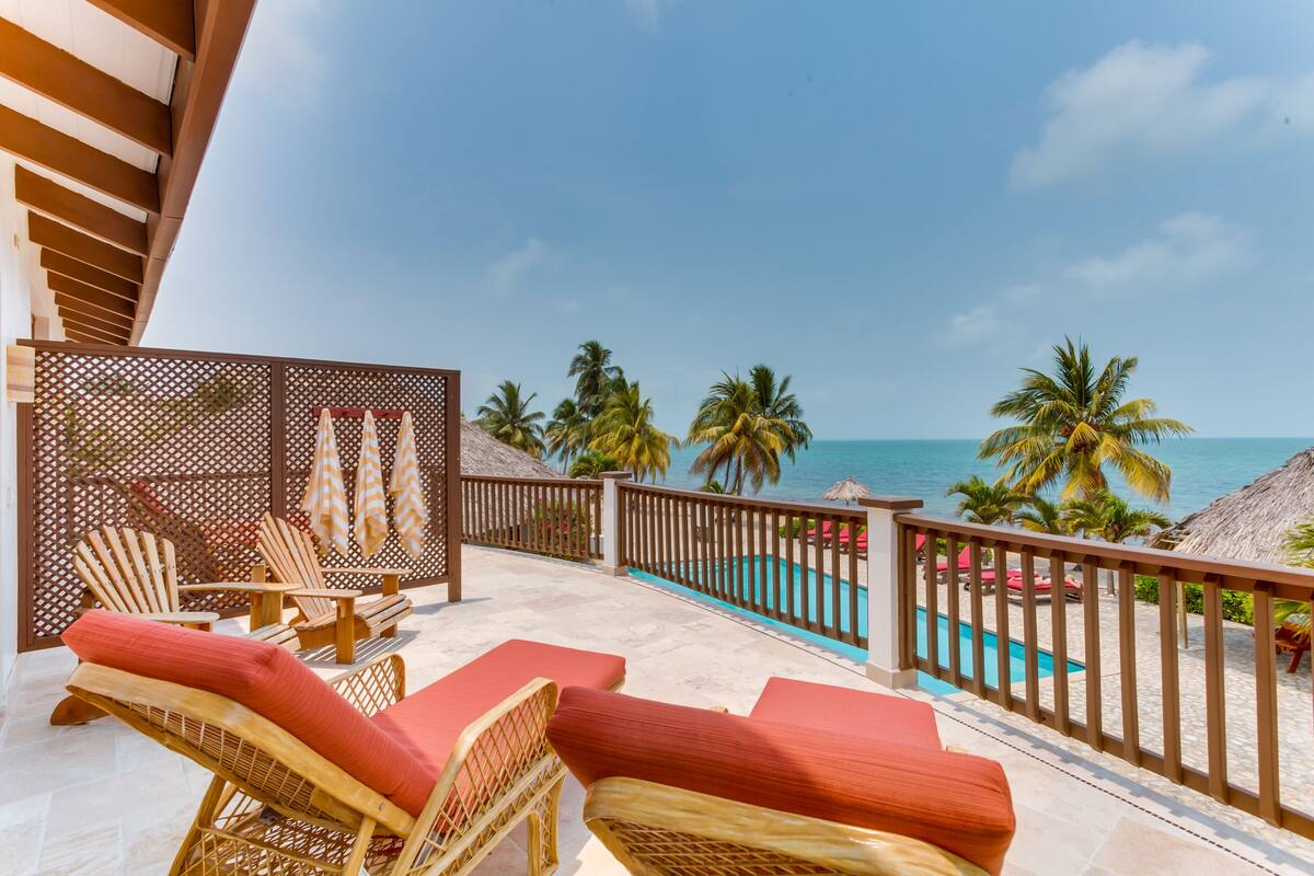 A balcony with lounge chairs and a pool, overlooking the ocean and palm trees.