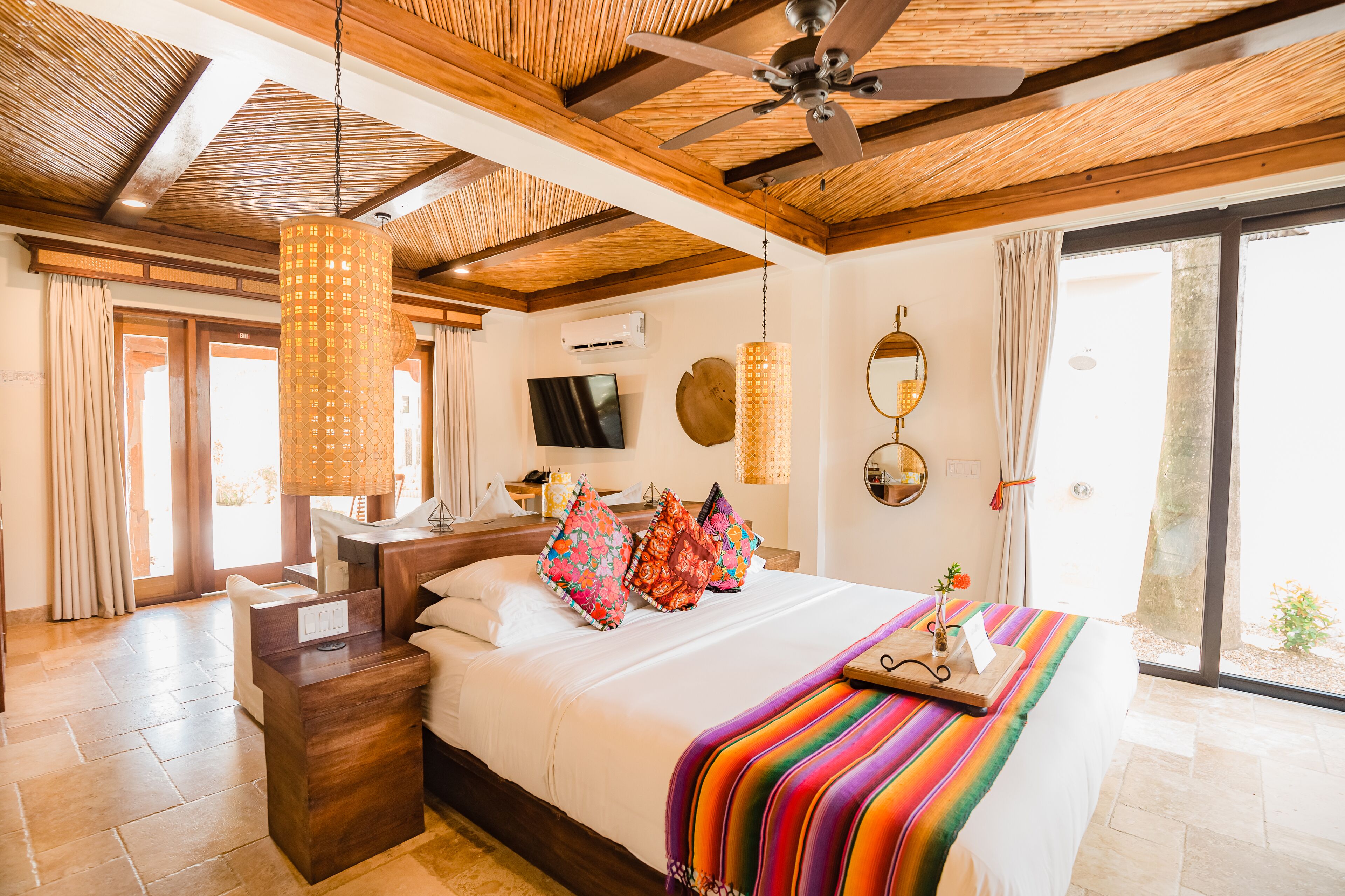 Une chambre d’hôtel avec un plafond en bois, un lit agrémenté d’oreillers colorés, une table de chevet, une télévision et une grande fenêtre.