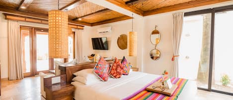 A hotel room with a wooden ceiling, a bed with colorful pillows, a bedside table, a TV, and a large window.