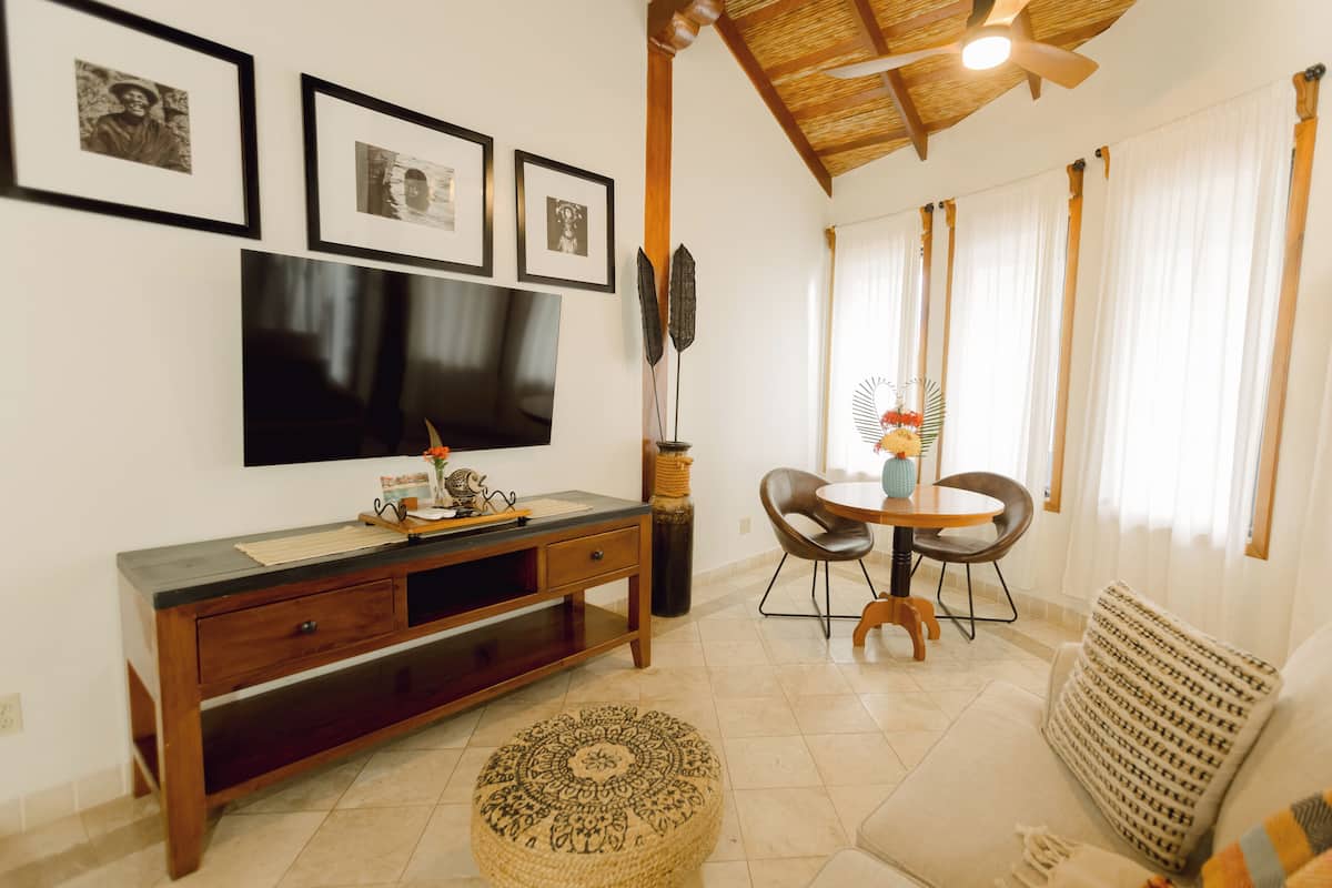 A living room with a TV mounted on the wall, a wooden console table, a round dining table with chairs, and a patterned ottoman.