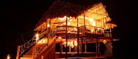 A multi-level wooden structure with a thatched roof, illuminated by warm lighting, and connected to a pool area.