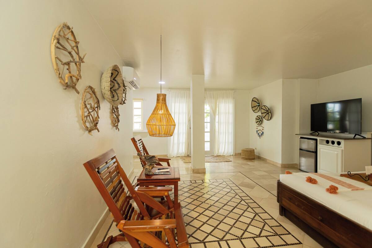 A hotel room with a bed, a TV, a dining table with chairs, a kitchen area, and decorative wall art.