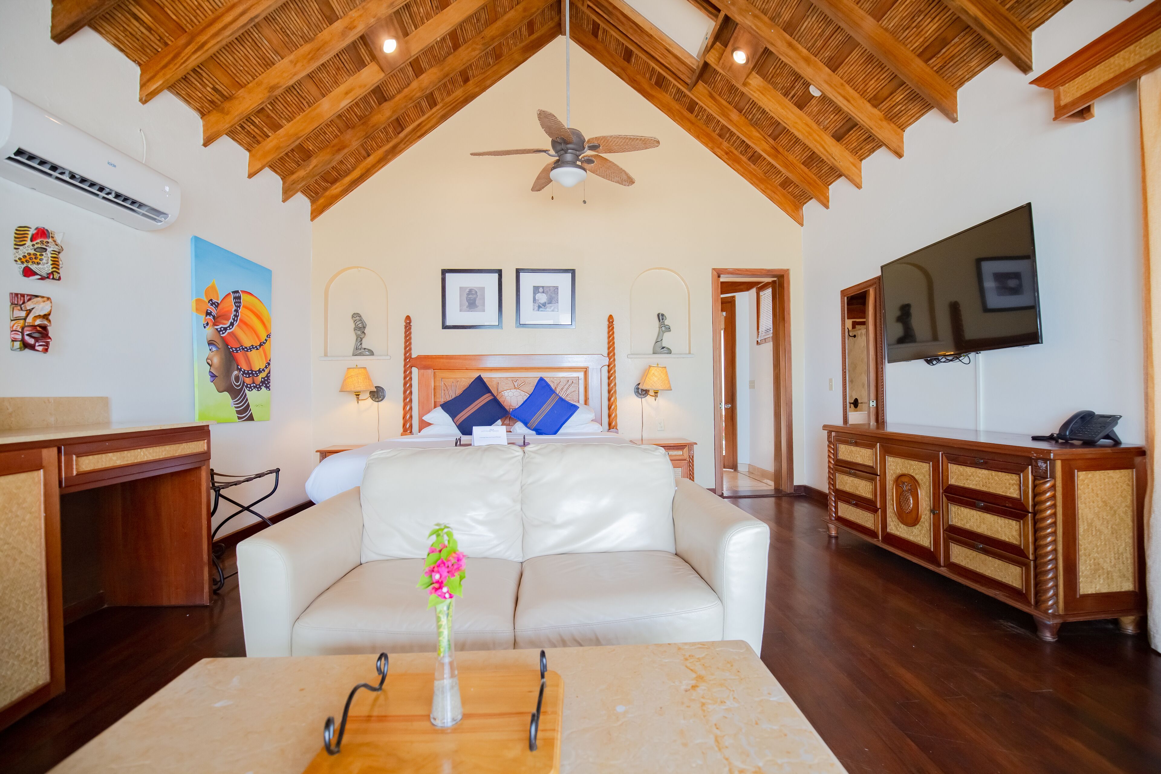 Uma sala de estar espaçosa com teto alto, ventilador de teto, uma cama grande com almofadas decorativas, uma TV e um armário de madeira.