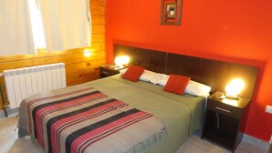 A bedroom with a wooden headboard, a green bedspread, two pillows, a nightstand with a lamp, and a radiator. - Hotel Punta Norte (El Calafate)