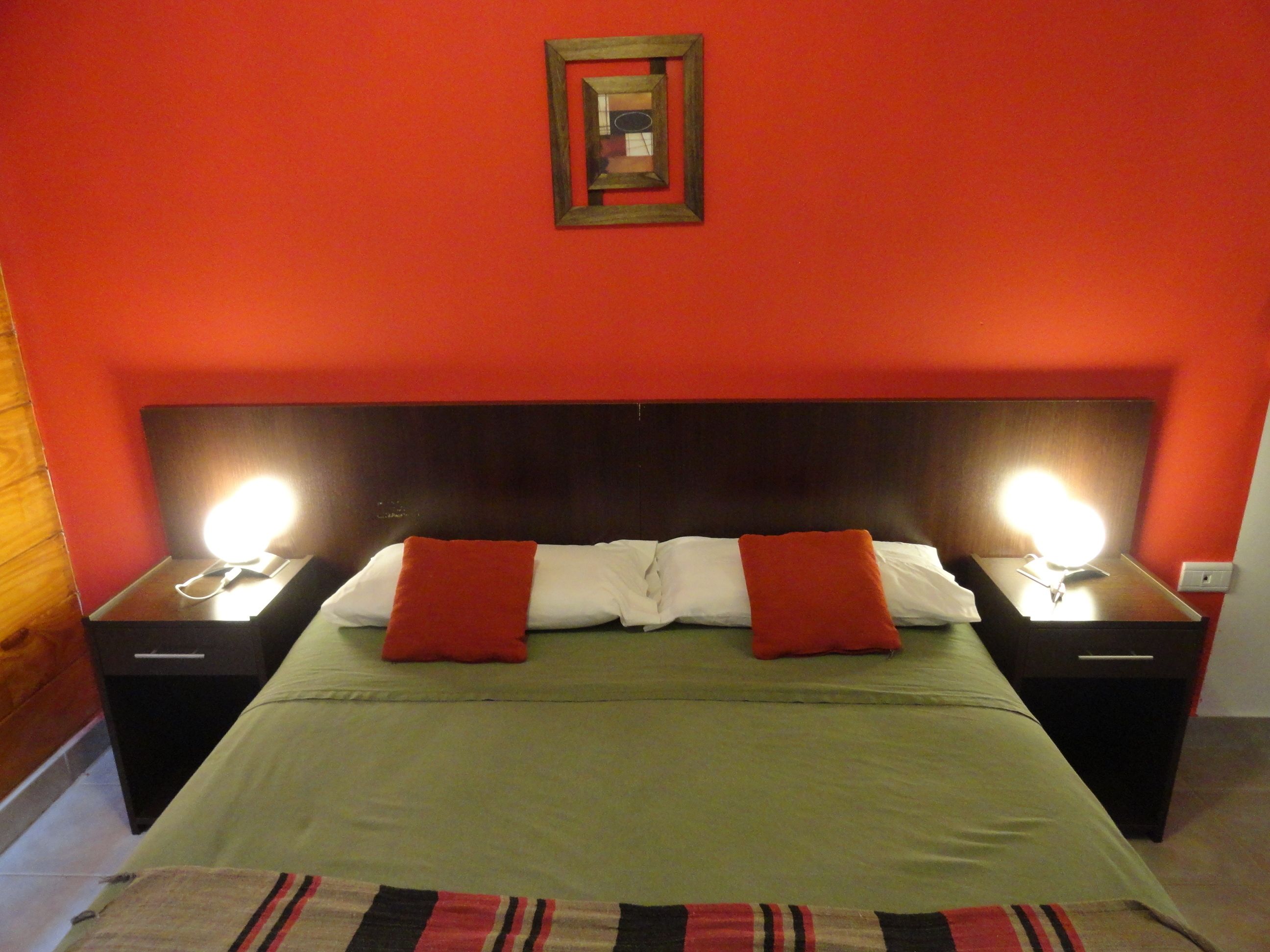 A hotel room with a large bed, two bedside tables with lamps, a red wall, and a framed picture above the bed.