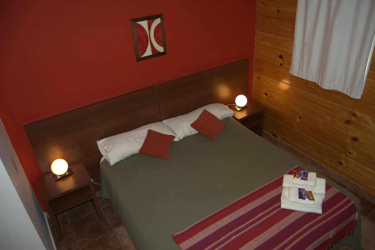 A single bed with white and red pillows, a striped blanket, and a small table with a lamp.