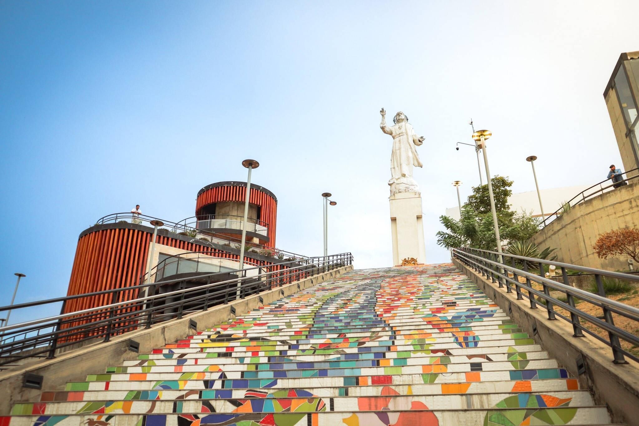 Eine Treppe mit bunten Fliesen führt zu einer Statue, ein rotes, zylinderförmiges Gebäude und ein klarer Himmel.