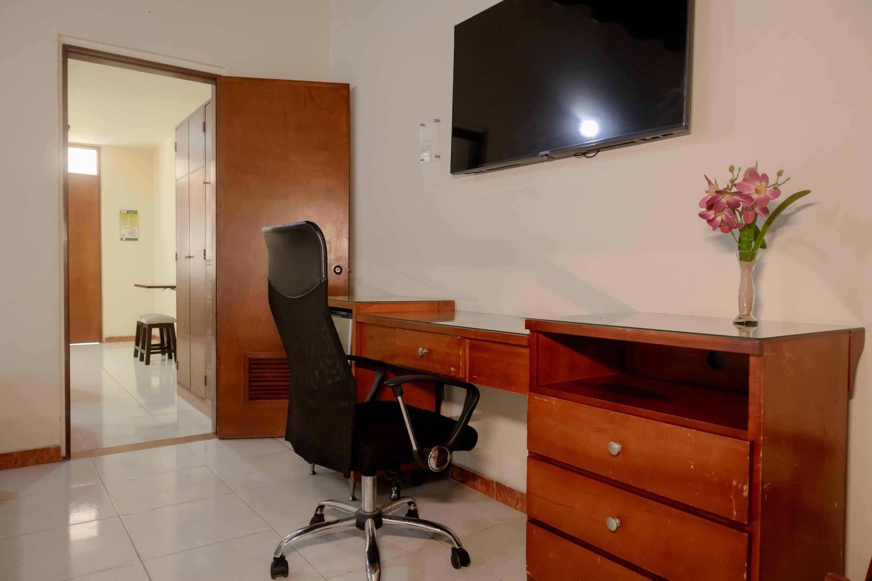 Una habitación con un escritorio de madera, una silla de oficina negra, un televisor montado en la pared y un jarrón pequeño con flores.