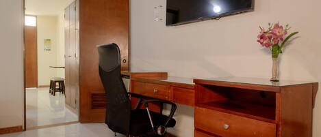 Una habitación con un escritorio de madera, una silla de oficina negra, un televisor montado en la pared y un jarrón pequeño con flores.