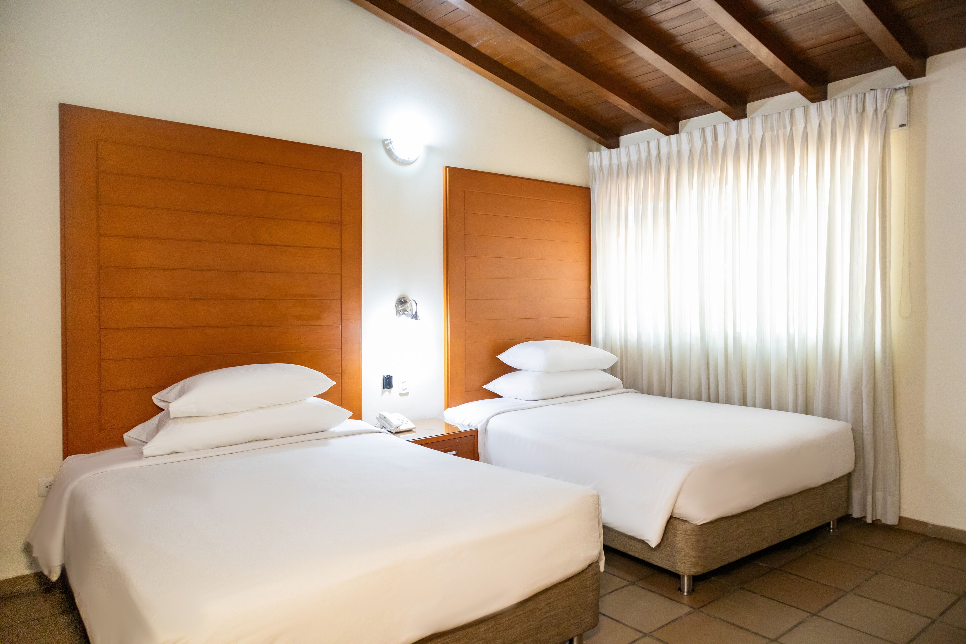 Habitación de hotel con dos camas, cabeceros de madera, una mesita de noche con teléfono y una ventana con cortinas.
