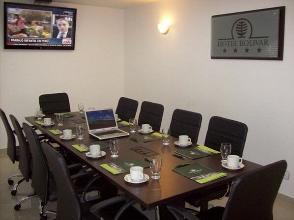 A conference room with a long table, black chairs, and a laptop. There are water glasses and cups on the table. A TV screen displays a news program. A sign for Hotel Bolívar is on the wall.