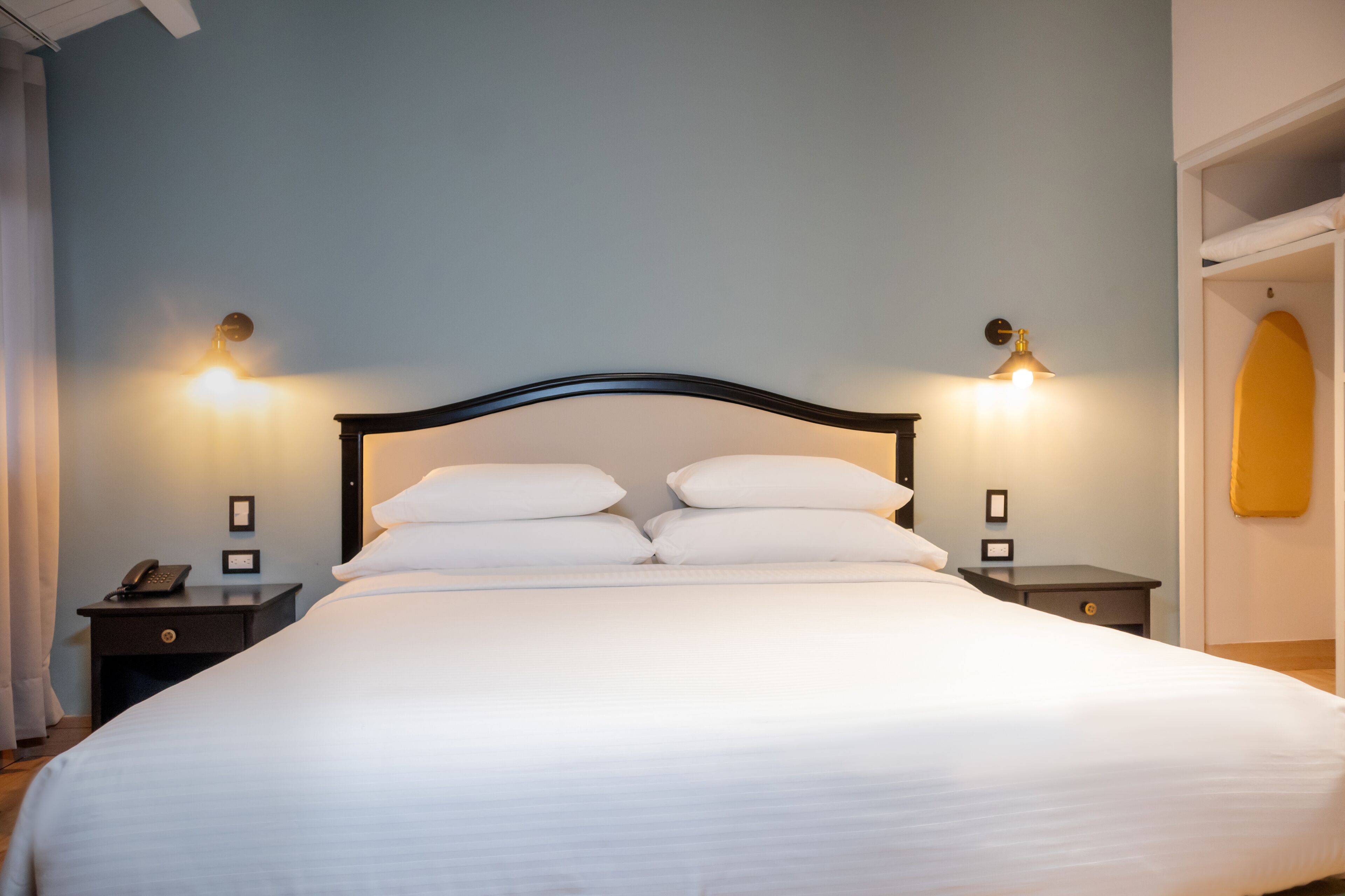 Cama bem arrumada com lençóis brancos e cabeceira de madeira. Duas mesas de cabeceira, com abajures e um telefone em uma delas. As paredes são pintadas de azul claro, e há uma prancha de surf na parede à direita.