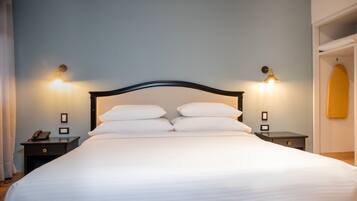 Cama bem arrumada com lençóis brancos e cabeceira de madeira. Duas mesas de cabeceira, com abajures e um telefone em uma delas. As paredes são pintadas de azul claro, e há uma prancha de surf na parede à direita.