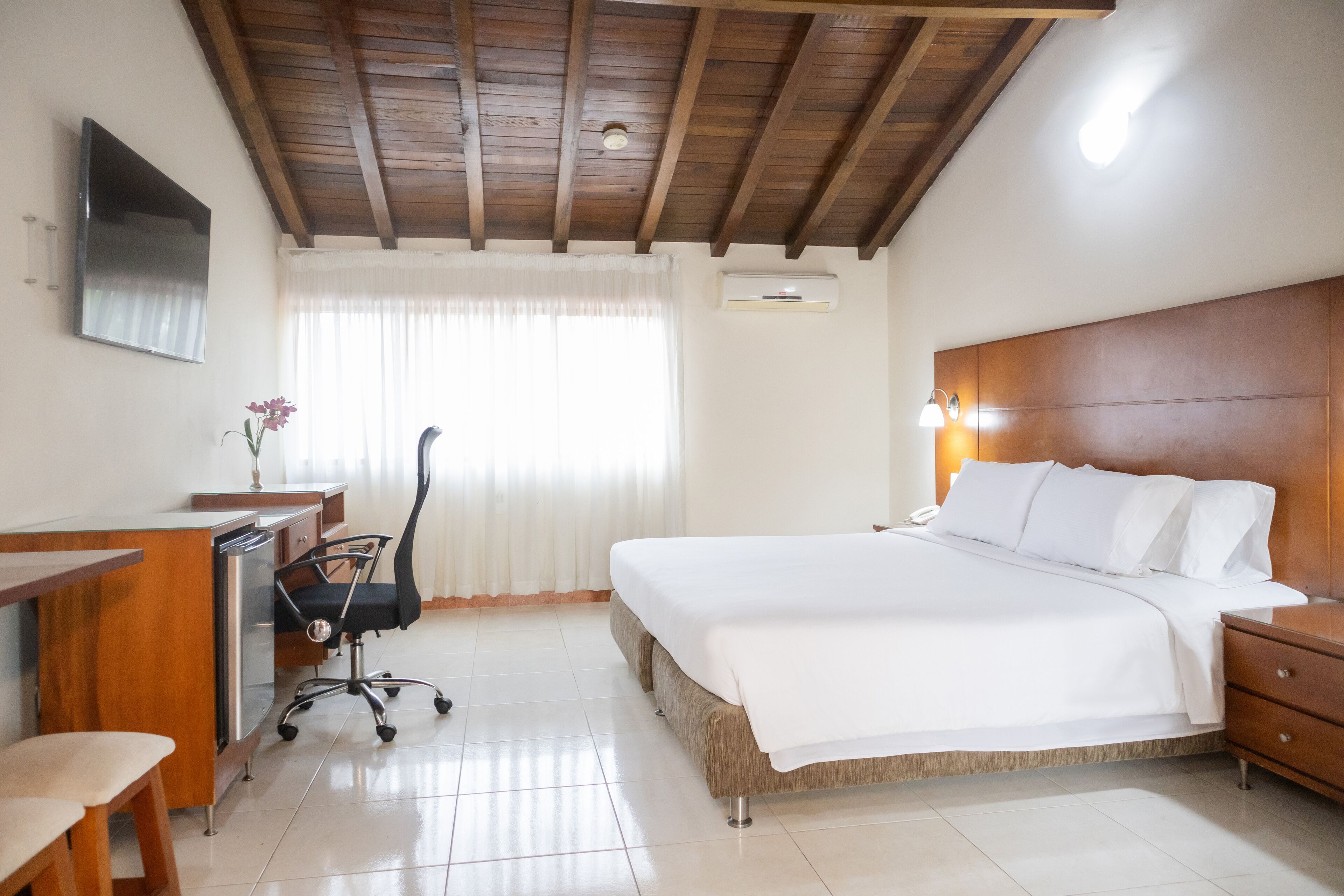 Une chambre avec un plafond en bois, un lit avec une literie blanche, un bureau avec une chaise et une fenêtre avec des rideaux.
