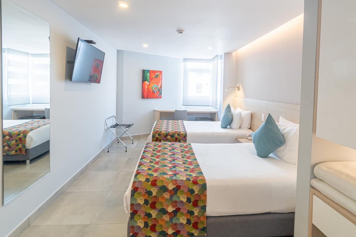Habitación de hotel con dos camas, un televisor montado en la pared, un escritorio y una ventana.