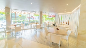 A modern indoor dining area with white tables and chairs, large windows, and a view of the street outside.