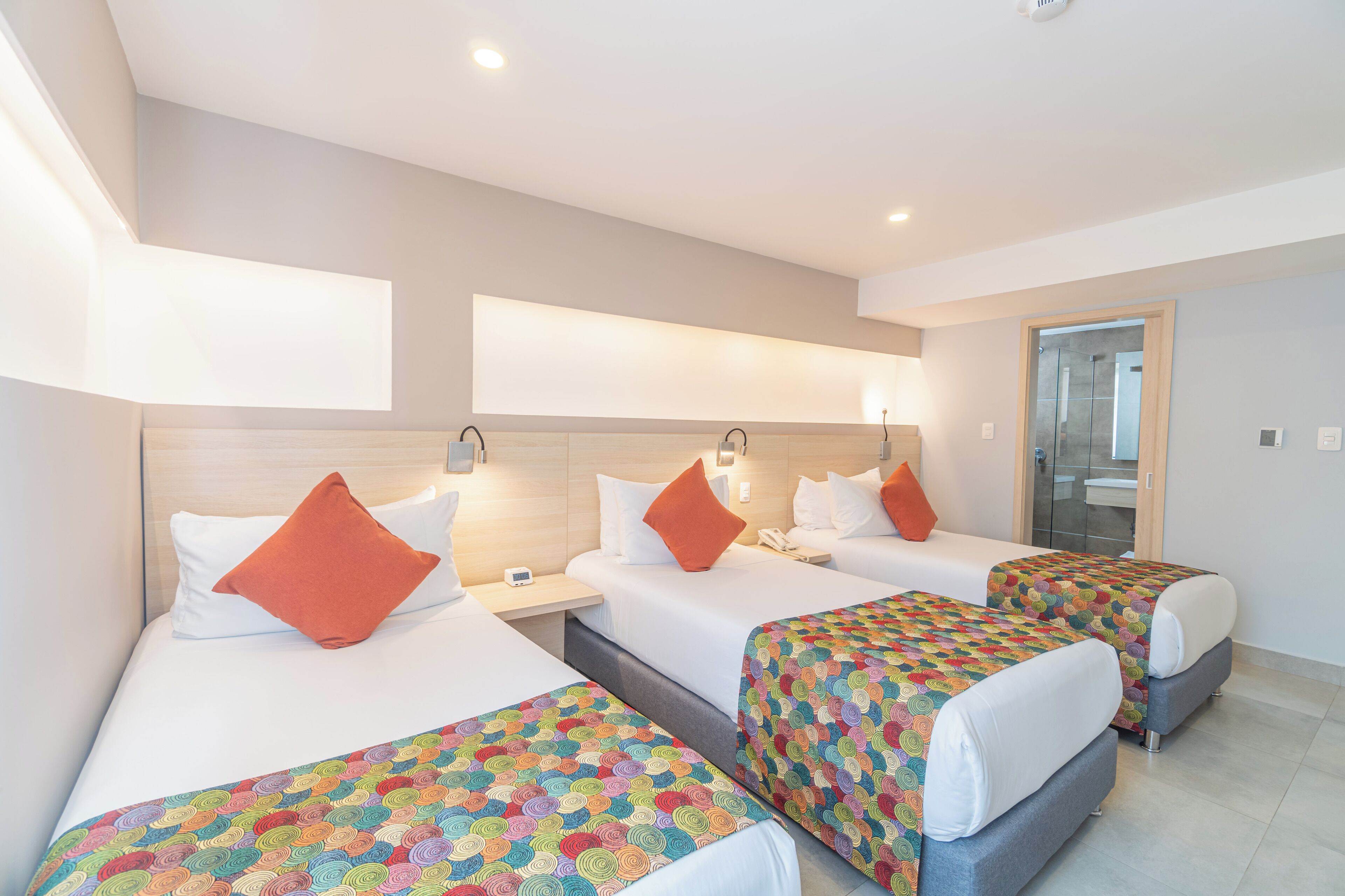 Ein Hotelzimmer mit drei Betten, jeweils mit einer farbenfrohen, gemusterten Tagesdecke und weißen Kissen.