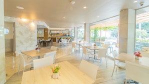 A spacious restaurant with multiple tables and chairs, a kitchen area in the background, and large windows offering views of the outdoors.