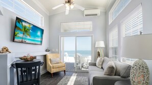 Uma sala de estar moderna com uma televisão grande, um sofá confortável, uma área de jantar com mesa e cadeiras, e vista para o oceano através de janelas amplas.