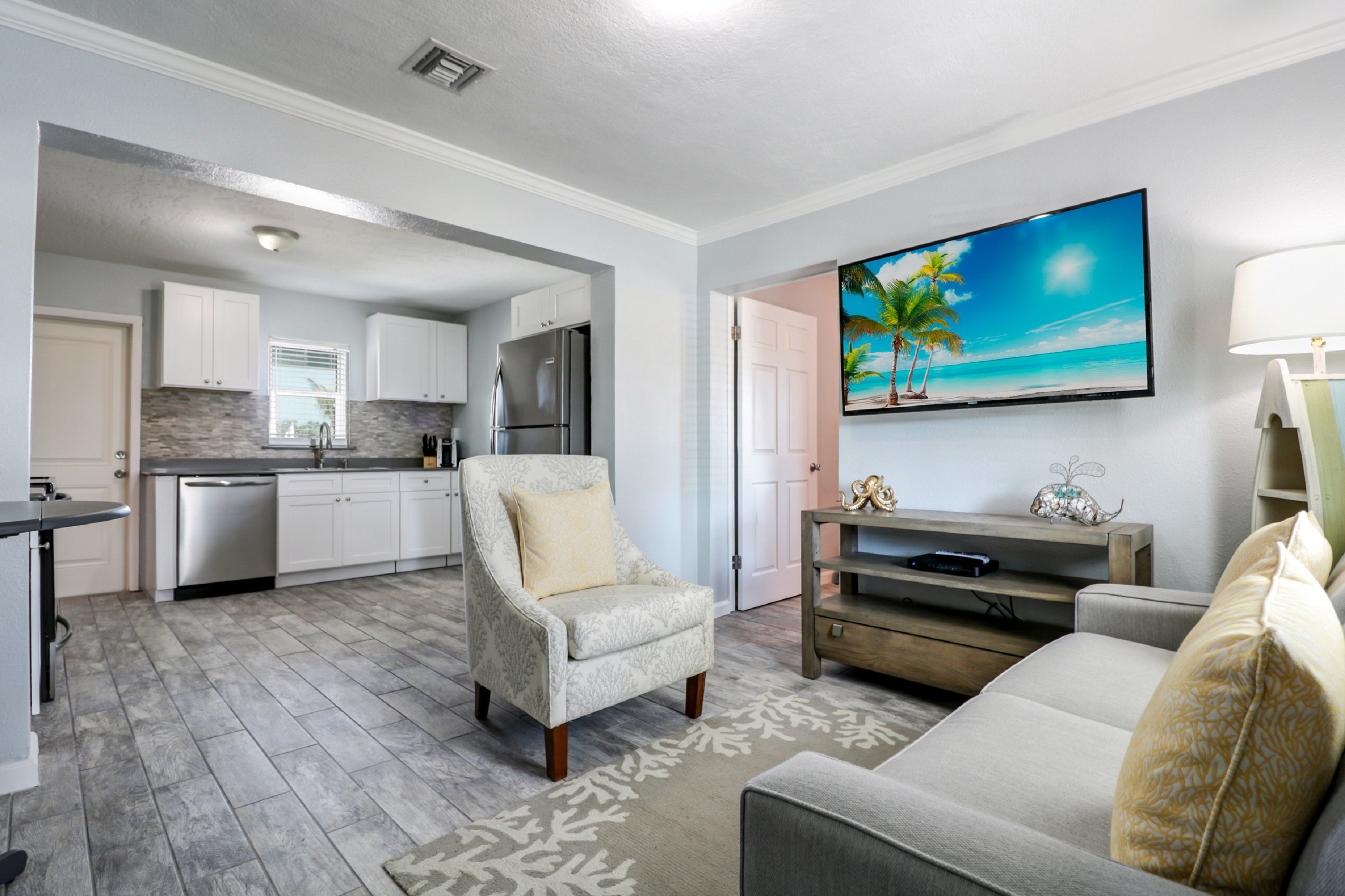 A modern kitchen with white cabinets, a stainless steel refrigerator, and a dishwasher. There is a dining area with a gray sofa and a patterned rug. A flat-screen TV is mounted on the wall above the sofa.