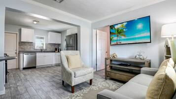 A modern kitchen with white cabinets, a stainless steel refrigerator, and a dishwasher. There is a dining area with a gray sofa and a patterned rug. A flat-screen TV is mounted on the wall above the sofa.