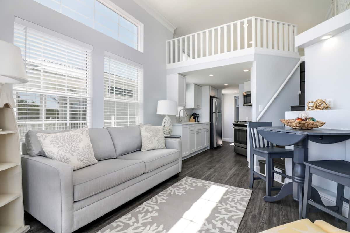 A modern kitchen with white cabinets, a dining area with a table and chairs, and a gray sofa with patterned cushions.