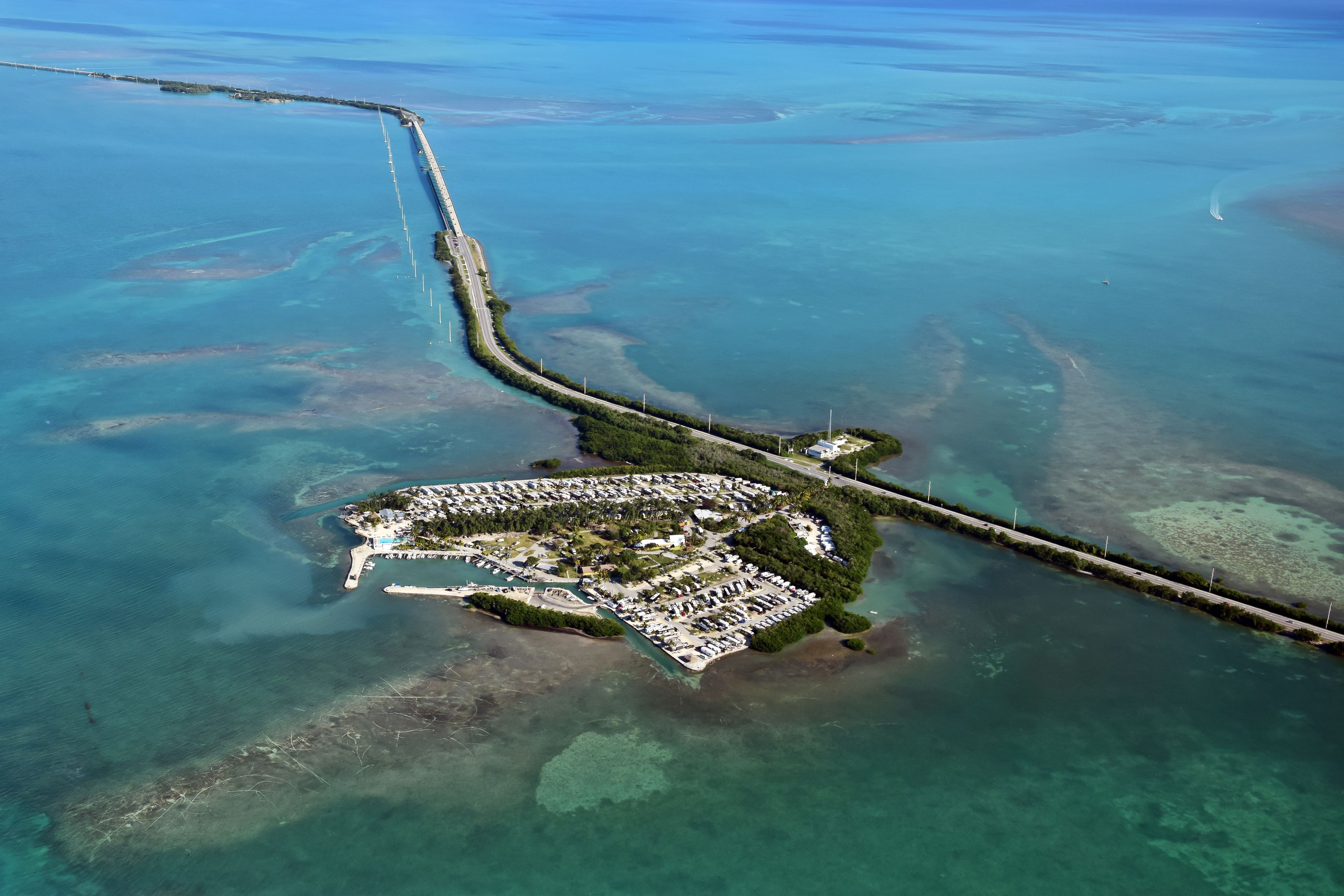 An aerial view of a coastal resort with a long pier extending into the sea, surrounded by clear blue waters and a well-organized marina.