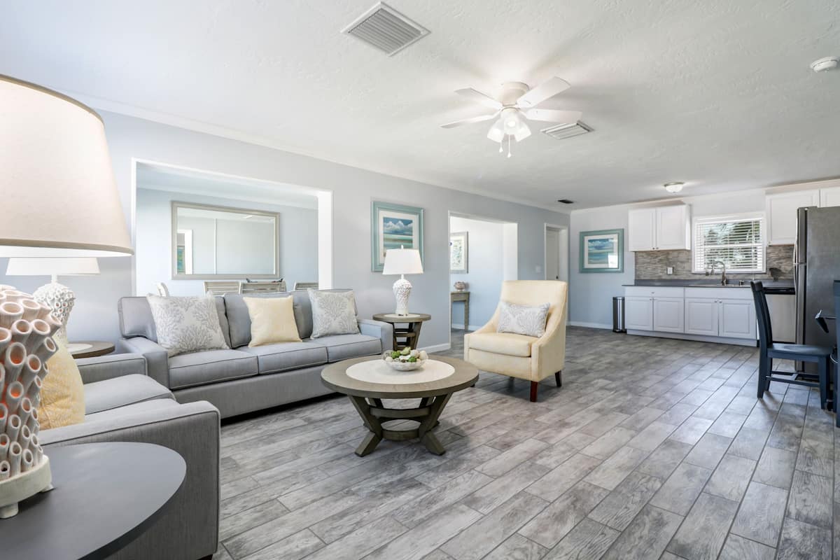 A spacious living room with a grey sofa, a round coffee table, and a chair. The kitchen area is visible with white cabinets and a refrigerator.