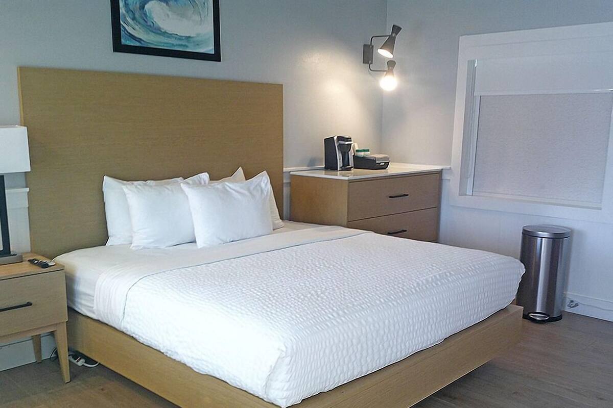 A neatly made bed with white linens, a wooden headboard, a bedside table with a lamp, and a trash bin.
