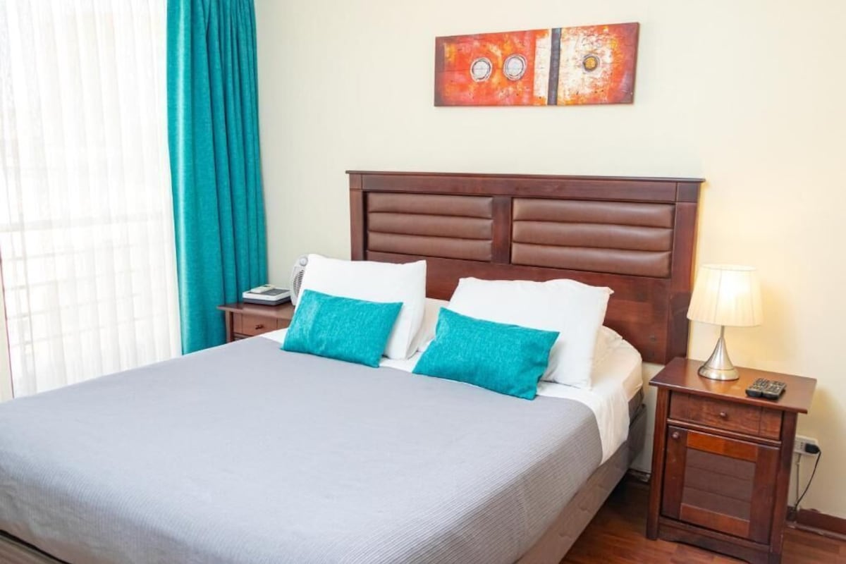 Un lit bien fait, recouvert d’une couvre-lit grise et agrémenté de deux coussins turquoise, avec une tête de lit en bois, une table de chevet dotée d’une lampe et une œuvre d’art murale au-dessus du lit.